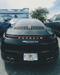 A black Porsche 911 Carrera 4S is parked between two other vehicles in an outdoor setting. The car has a Florida license plate and is positioned in front of a mural depicting ocean scenes with dolphins and stingrays. The background includes palm trees and a partly cloudy sky.