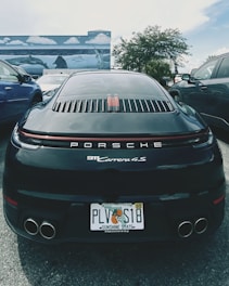 A black Porsche 911 Carrera 4S is parked between two other vehicles in an outdoor setting. The car has a Florida license plate and is positioned in front of a mural depicting ocean scenes with dolphins and stingrays. The background includes palm trees and a partly cloudy sky.