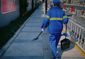 A person wearing a blue uniform with reflective stripes is walking on a paved sidewalk. They are carrying a dustpan and a brush. Along the side of the walkway, there are potted plants and a fence. In the background, there are more people and flower arrangements.