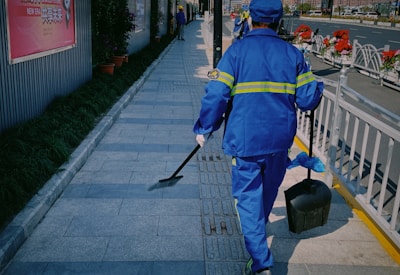 A person wearing a blue uniform with reflective stripes is walking on a paved sidewalk. They are carrying a dustpan and a brush. Along the side of the walkway, there are potted plants and a fence. In the background, there are more people and flower arrangements.