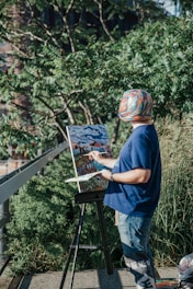 A person wearing a colorful hat and a blue shirt is painting on a canvas set up on an easel outdoors. The person is surrounded by lush green foliage and the scene appears to be sunny. The painting they are working on depicts a landscape with a bridge and buildings under a cloudy sky.