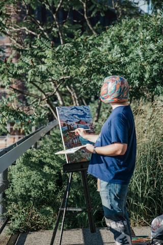 A person wearing a colorful hat and a blue shirt is painting on a canvas set up on an easel outdoors. The person is surrounded by lush green foliage and the scene appears to be sunny. The painting they are working on depicts a landscape with a bridge and buildings under a cloudy sky.