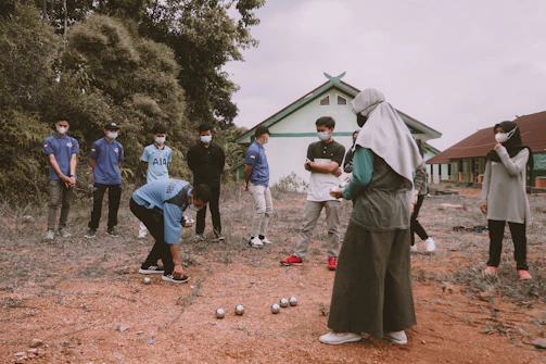 A group of people, mostly wearing masks, is gathered outdoors. Several are standing back, observing, while one person is bending down, possibly engaged in a game involving small metallic balls laid out on the ground. The setting includes a building with a green roof in the background and some vegetation around.