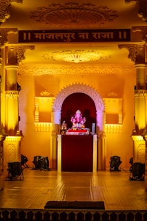 An ornate stage decorated with intricate carvings and arches, illuminated by warm yellow lighting. At the center of the stage is a small idol of Lord Ganesha, adorned with garlands and surrounded by religious offerings.
