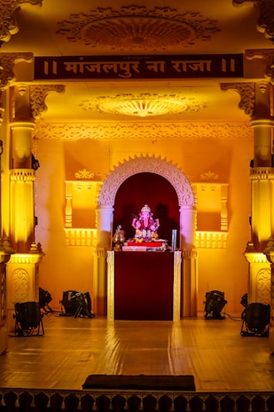 An ornate stage decorated with intricate carvings and arches, illuminated by warm yellow lighting. At the center of the stage is a small idol of Lord Ganesha, adorned with garlands and surrounded by religious offerings.