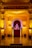 An ornate stage decorated with intricate carvings and arches, illuminated by warm yellow lighting. At the center of the stage is a small idol of Lord Ganesha, adorned with garlands and surrounded by religious offerings.