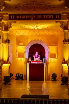 An ornate stage decorated with intricate carvings and arches, illuminated by warm yellow lighting. At the center of the stage is a small idol of Lord Ganesha, adorned with garlands and surrounded by religious offerings.
