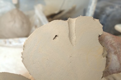 A hand is holding a piece of light-colored clay or dough. The surface displays a rough texture with a small hole and a notch at the edge. The background appears blurred, suggesting a workshop or studio environment.
