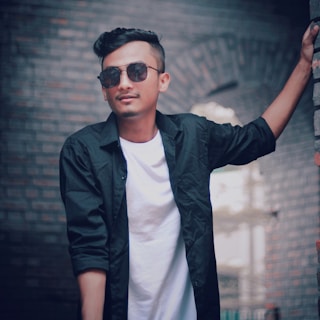 A man dressed in a sleek black kixoraa shirt, leaning against a brick wall with a relaxed pose.