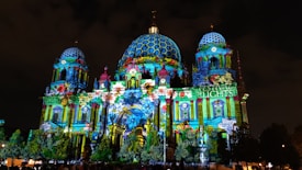 A grand building is illuminated with vibrant, colorful projections featuring intricate patterns and designs. The projections cover the entire facade of the structure, adding a lively and festive atmosphere. The words 'Festival of Lights' are prominently displayed on one section of the building.