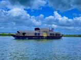 A traditional houseboat with woven wicker cabins sails gently across a serene body of water under a partly cloudy sky. The boat is equipped with amenities and several people are visible on the deck, enjoying the view surrounded by lush green scenery.