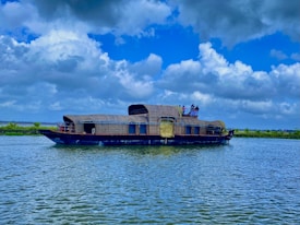 A traditional houseboat with woven wicker cabins sails gently across a serene body of water under a partly cloudy sky. The boat is equipped with amenities and several people are visible on the deck, enjoying the view surrounded by lush green scenery.