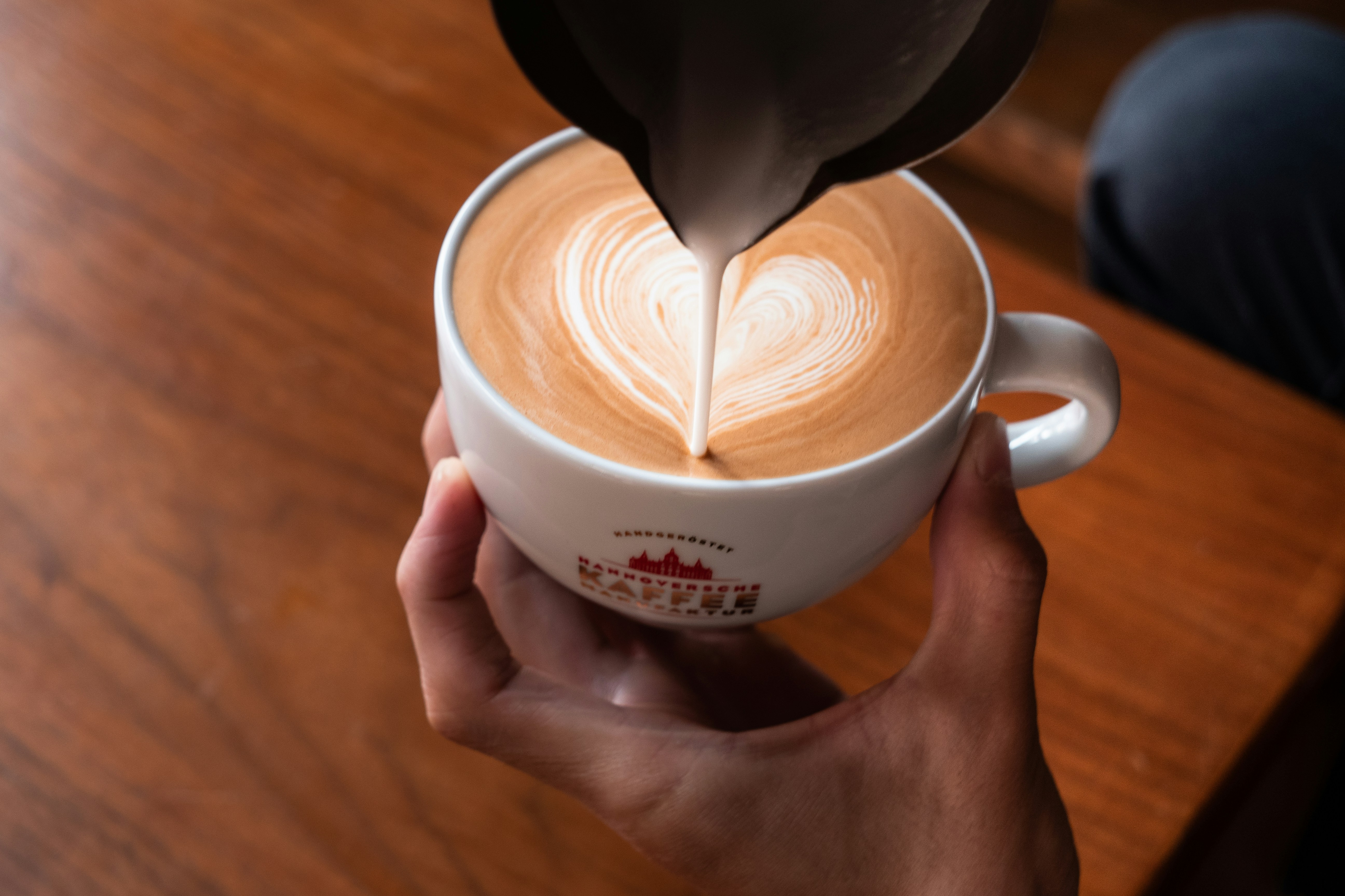 Latte art heart being poured into ceramic cup, white steamed milk swirling into dark espresso, bright airy café window light
