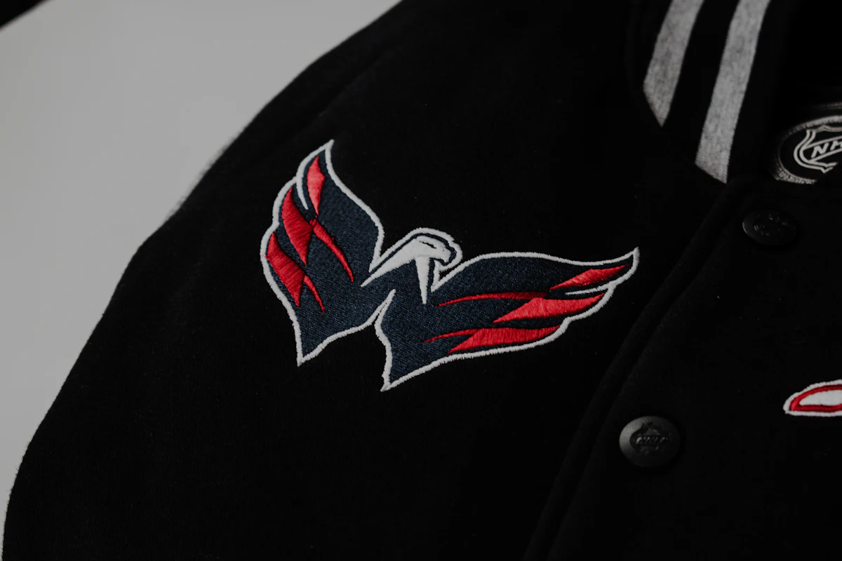 Jaket varsity hitam dilihat dari depan miring: deretan kancing snap di bagian tengah, kerah rib bergaris, dan patch bordir berjahit keliling di dada.