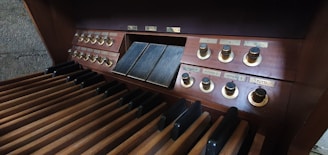 A close-up of a wooden organ console featuring multiple foot pedals and control knobs. The pedals are aligned in rows, while the control panel has labels and buttons arranged in sections. The wood finish gives a vintage look to the instrument.