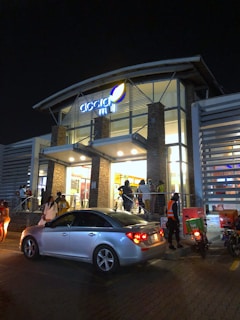 A modern building with glass walls is illuminated at night. Several people are gathered near the entrance, which has bright lights and welcoming signs. A silver sedan is parked in front of the building, while two motorbikes are nearby, one with a food delivery box.