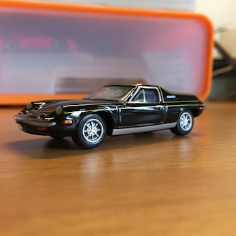 A small-scale model of a black sports car is placed on a wooden surface. The car has detailed features and shiny chrome wheels. In the background, there is a blurred orange and translucent container.