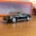 A small-scale model of a black sports car is placed on a wooden surface. The car has detailed features and shiny chrome wheels. In the background, there is a blurred orange and translucent container.