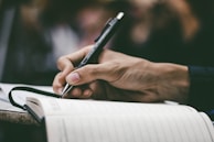 A hand is holding a pen and writing in a notebook. The scene is focused on the act of writing, with a close-up view that captures the details of the hand and pen. The background is blurred, emphasizing the notebook and hand.