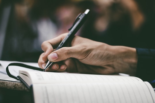 A hand is holding a pen and writing in a notebook. The scene is focused on the act of writing, with a close-up view that captures the details of the hand and pen. The background is blurred, emphasizing the notebook and hand.