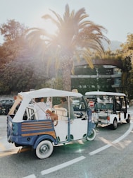 A bright and sunny scene featuring a palm tree prominently in the background. Two three-wheeler tuk-tuks are on a curved road, with the sunlight creating a flare effect. People are inside the tuk-tuks, which have an open-air design. Surrounding greenery and a partial view of a building add to the relaxed, tropical atmosphere.