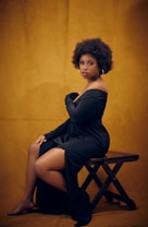 A woman with afro-textured hair is seated on a wooden stool, wearing a long, off-the-shoulder black dress with a thigh-high slit. The background is a rich, mustard yellow color, adding warmth to the composition. She is looking directly at the camera with a calm and confident expression.