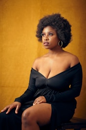 A woman with an afro hairstyle sits against a warm yellow background, wearing a black off-the-shoulder dress and hoop earrings. Her expression is calm and contemplative, with her hands resting on her lap.