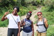 Three women are standing on a dirt path surrounded by lush greenery, smiling and holding up peace signs. They are dressed casually, wearing athletic and comfortable clothing suitable for hiking. Each of them is wearing sunglasses, and two have red bandanas.