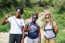 Three women are standing on a dirt path surrounded by lush greenery, smiling and holding up peace signs. They are dressed casually, wearing athletic and comfortable clothing suitable for hiking. Each of them is wearing sunglasses, and two have red bandanas.