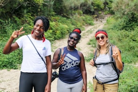 Three women are standing on a dirt path surrounded by lush greenery, smiling and holding up peace signs. They are dressed casually, wearing athletic and comfortable clothing suitable for hiking. Each of them is wearing sunglasses, and two have red bandanas.