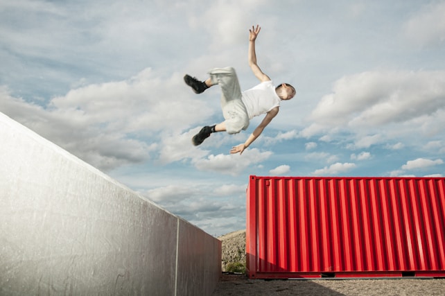 A person is performing a dynamic flip or jump between two large structures. The individual is mid-air, with arms extended and legs bent. One structure is metallic and silver, while the other is a vibrant red container. The sky is expansive, filled with light clouds.