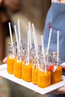A set of nine small glass bottles filled with a bright orange liquid, each topped with a white straw, are arranged neatly on a white tray. The bottles have metal swing-top closures and are being carried by a person wearing a blue apron.