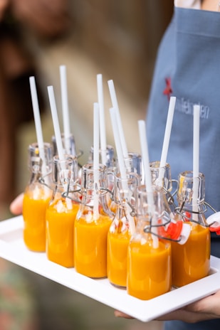 A set of nine small glass bottles filled with a bright orange liquid, each topped with a white straw, are arranged neatly on a white tray. The bottles have metal swing-top closures and are being carried by a person wearing a blue apron.