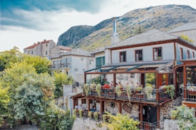 A picturesque outdoor restaurant terrace set in a rustic building with stone and wood elements, surrounded by lush greenery and mountainous landscape. Diners sit at tables adorned with vibrant flowers, enjoying a peaceful atmosphere.