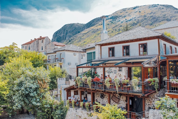 A picturesque outdoor restaurant terrace set in a rustic building with stone and wood elements, surrounded by lush greenery and mountainous landscape. Diners sit at tables adorned with vibrant flowers, enjoying a peaceful atmosphere.