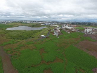 A sprawling landscape with lush green fields dominates the foreground, leading towards a body of water surrounded by vegetation. In the distance, a collection of modern buildings under construction is visible, with cranes scattered throughout. The sky is overcast with thick clouds, casting a dull light over the scene.