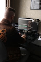 A person with a distinct hairstyle sits at a desk, working on two computer monitors displaying code. The person wears a patterned sweater with animal motifs. The room has dim lighting with a warm color palette, and there is a framed poster on the wall.
