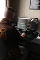 A person with a distinct hairstyle sits at a desk, working on two computer monitors displaying code. The person wears a patterned sweater with animal motifs. The room has dim lighting with a warm color palette, and there is a framed poster on the wall.
