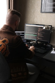 A person with a distinct hairstyle sits at a desk, working on two computer monitors displaying code. The person wears a patterned sweater with animal motifs. The room has dim lighting with a warm color palette, and there is a framed poster on the wall.