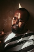 A person wearing glasses is holding a cigarette while smoke swirls around them. Soft, dramatic lighting creates shadows on the face and highlights the contours. The individual has a beard and is wearing a white shirt with bold black stripes, set against a neutral-colored background.