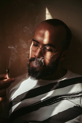 A person wearing glasses is holding a cigarette while smoke swirls around them. Soft, dramatic lighting creates shadows on the face and highlights the contours. The individual has a beard and is wearing a white shirt with bold black stripes, set against a neutral-colored background.