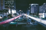 A nighttime urban cityscape featuring a busy road under construction, flanked by tall buildings with lit windows. Red and white streaks of light from vehicles create dynamic lines against the dark sky. Construction equipment and barriers are visible along the road.