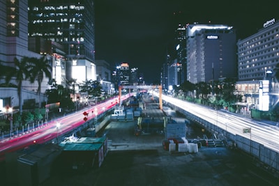 A nighttime urban cityscape featuring a busy road under construction, flanked by tall buildings with lit windows. Red and white streaks of light from vehicles create dynamic lines against the dark sky. Construction equipment and barriers are visible along the road.