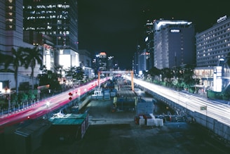 A nighttime urban cityscape featuring a busy road under construction, flanked by tall buildings with lit windows. Red and white streaks of light from vehicles create dynamic lines against the dark sky. Construction equipment and barriers are visible along the road.