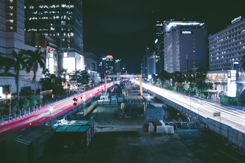 A nighttime urban cityscape featuring a busy road under construction, flanked by tall buildings with lit windows. Red and white streaks of light from vehicles create dynamic lines against the dark sky. Construction equipment and barriers are visible along the road.