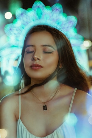 A woman with closed eyes is wearing a necklace and a light-colored dress, surrounded by a glow of circular lights creating a halo effect. The atmosphere is dreamy and ethereal with soft focus and blurred light patterns in the background.