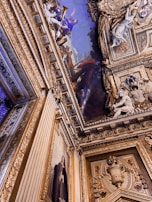 Ornate ceiling and wall with intricate gold and blue detailing. The ceiling features a vivid painting with classical figures, including one holding a trumpet. Decorative sculptures and frames add richness, and a large wall painting depicts a person in period clothing.