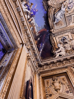 Ornate ceiling and wall with intricate gold and blue detailing. The ceiling features a vivid painting with classical figures, including one holding a trumpet. Decorative sculptures and frames add richness, and a large wall painting depicts a person in period clothing.