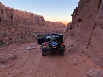 A rugged canyon landscape with tall, red rock cliffs on either side of a dusty, unpaved path. A black Jeep with its door open is parked on the path, surrounded by the striking geological formations that catch the warm hues of the setting sun.