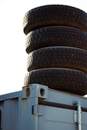 A stack of four dark, rugged tires is placed on top of a weathered metal container with visible rust stains and exposed metal hinges.